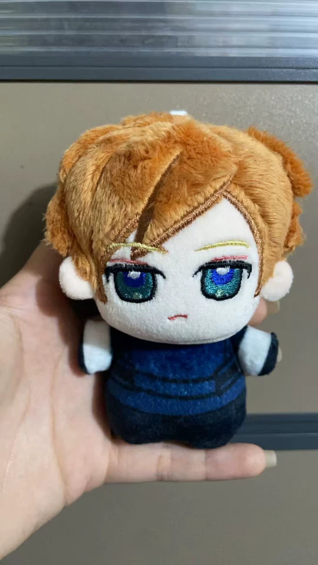 A small plush doll with orange-brown hair, large blue glittery eyes, and a blue outfit is being held in a person's hand against a gray indoor background.